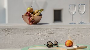 Una mesa con un cuenco de fruta, dos copas de vino y un libro en un balcón, con un edificio blanco al fondo y vista al mar.
