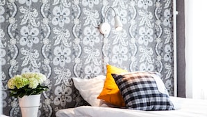 Quarto de hotel com papel de parede estampado, mesa de cabeceira com um vaso de flores e cama com almofadas brancas e amarelas.