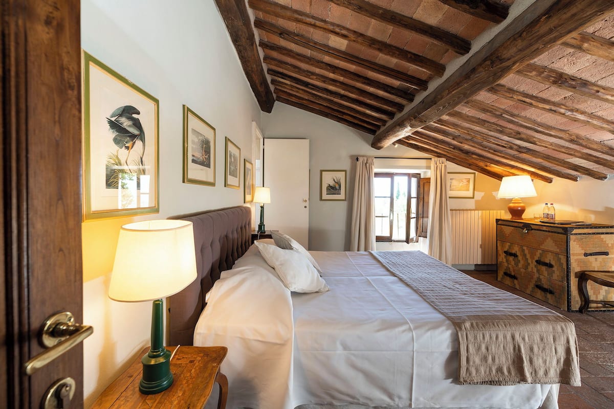 A bedroom with a wooden ceiling, a bed with white linens, a wooden nightstand with a lamp, and a dresser with a lamp.