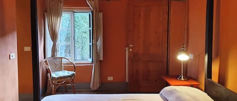 Un dormitorio con cama, una silla, una ventana con cortinas, una puerta de madera y una mesita de noche con lámpara.