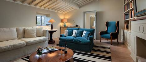 A living room with a wooden ceiling, a sofa, a blue sofa, a chair, a coffee table with wine glasses, and a bookshelf.