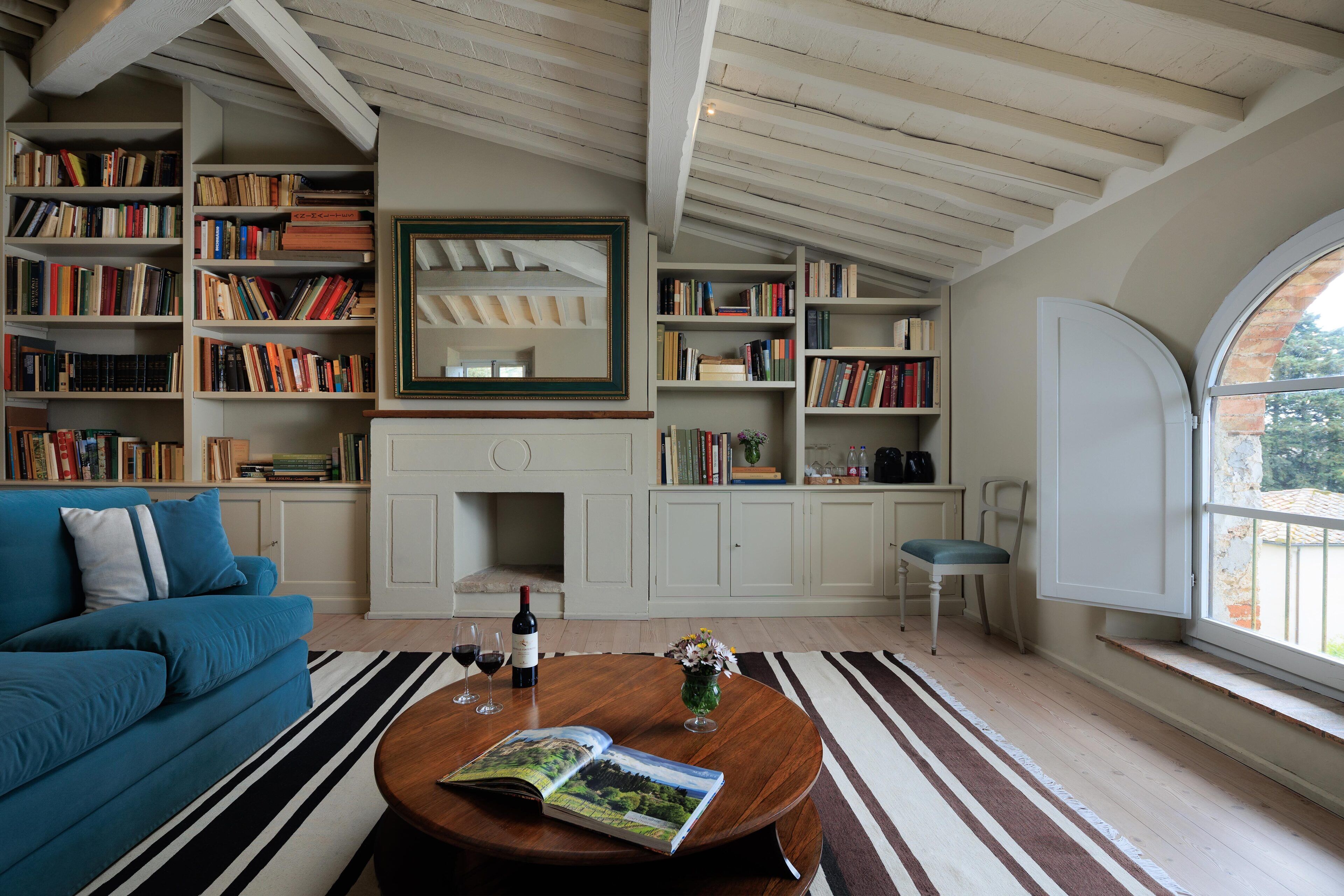 A cozy living room with a blue sofa, a wooden coffee table with a magazine, a bookshelf filled with books, a fireplace, and a large window with a view of greenery.