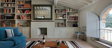 A cozy living room with a blue sofa, a wooden coffee table with a magazine, a bookshelf filled with books, a fireplace, and a large window with a view of greenery.