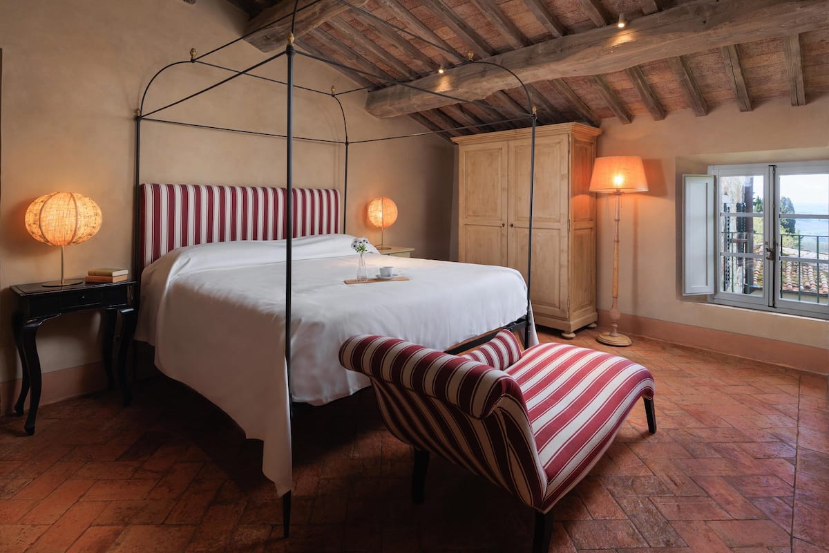 A four-poster bed with a canopy, a wooden wardrobe, a chair with striped upholstery, and a view of the sea through a window.