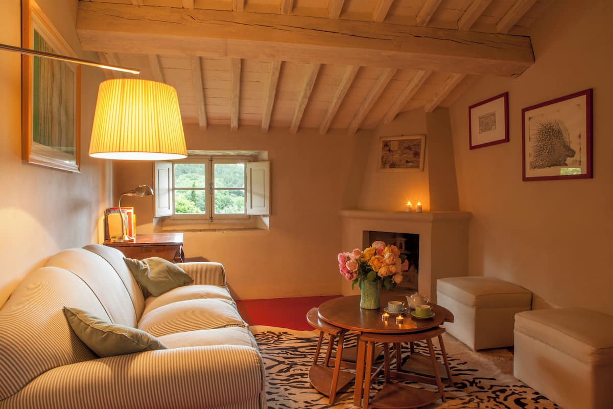 A cozy living room with a sofa, a small round table with a vase of flowers, a fireplace, and framed artwork on the walls.