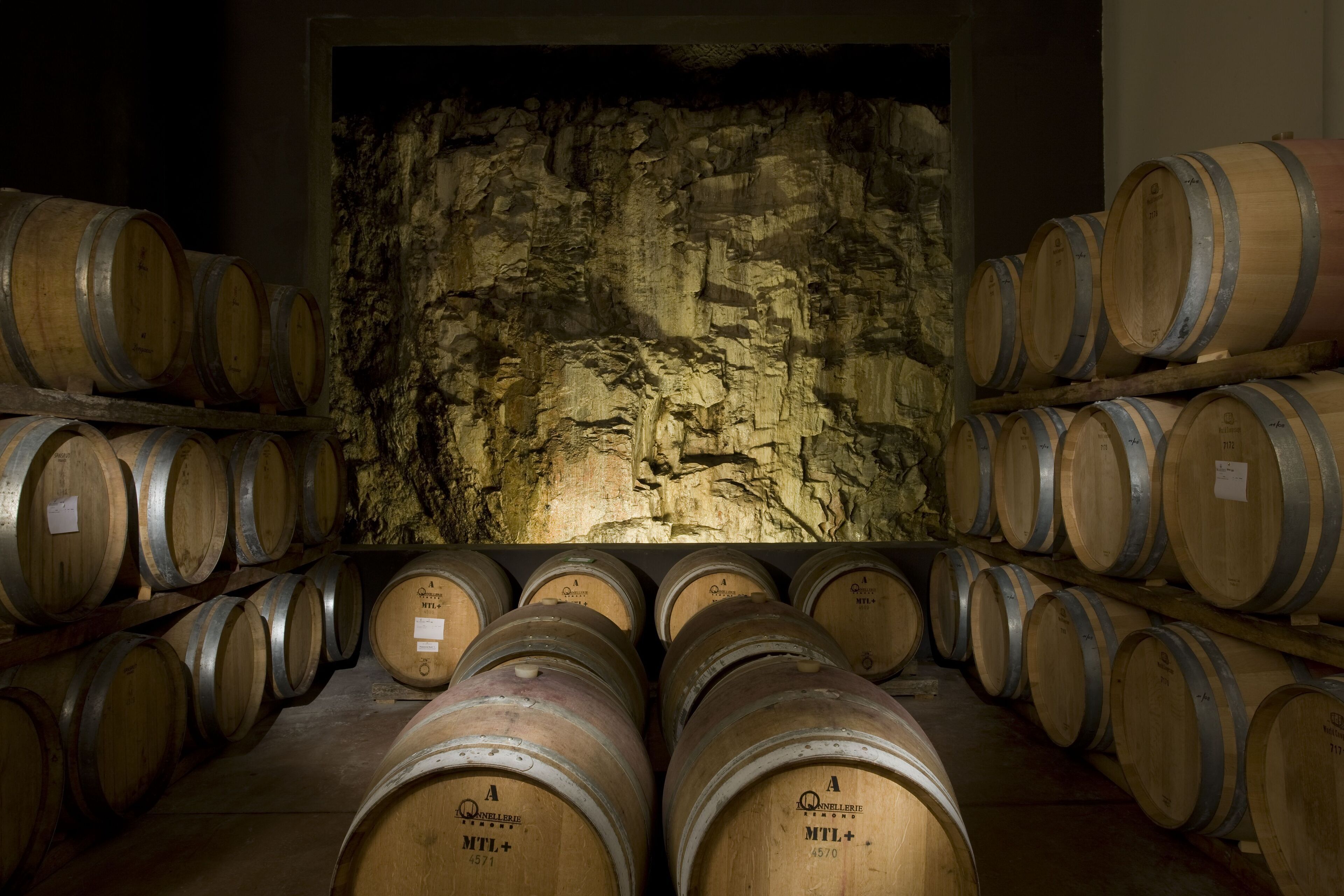 Wooden wine barrels stacked in a dimly lit cellar with a rough stone wall in the background.