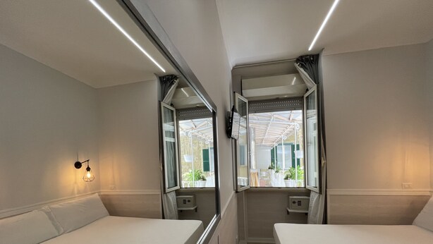 Un compartimento de tren con dos camas individuales, una mesita, una luz en la pared y una ventana que da a un balcón.