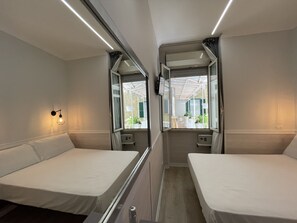 A train compartment with two single beds, a small table, a wall-mounted light, and a window with a view of an outdoor balcony. - Hotel Giotto Flavia (Rome)
