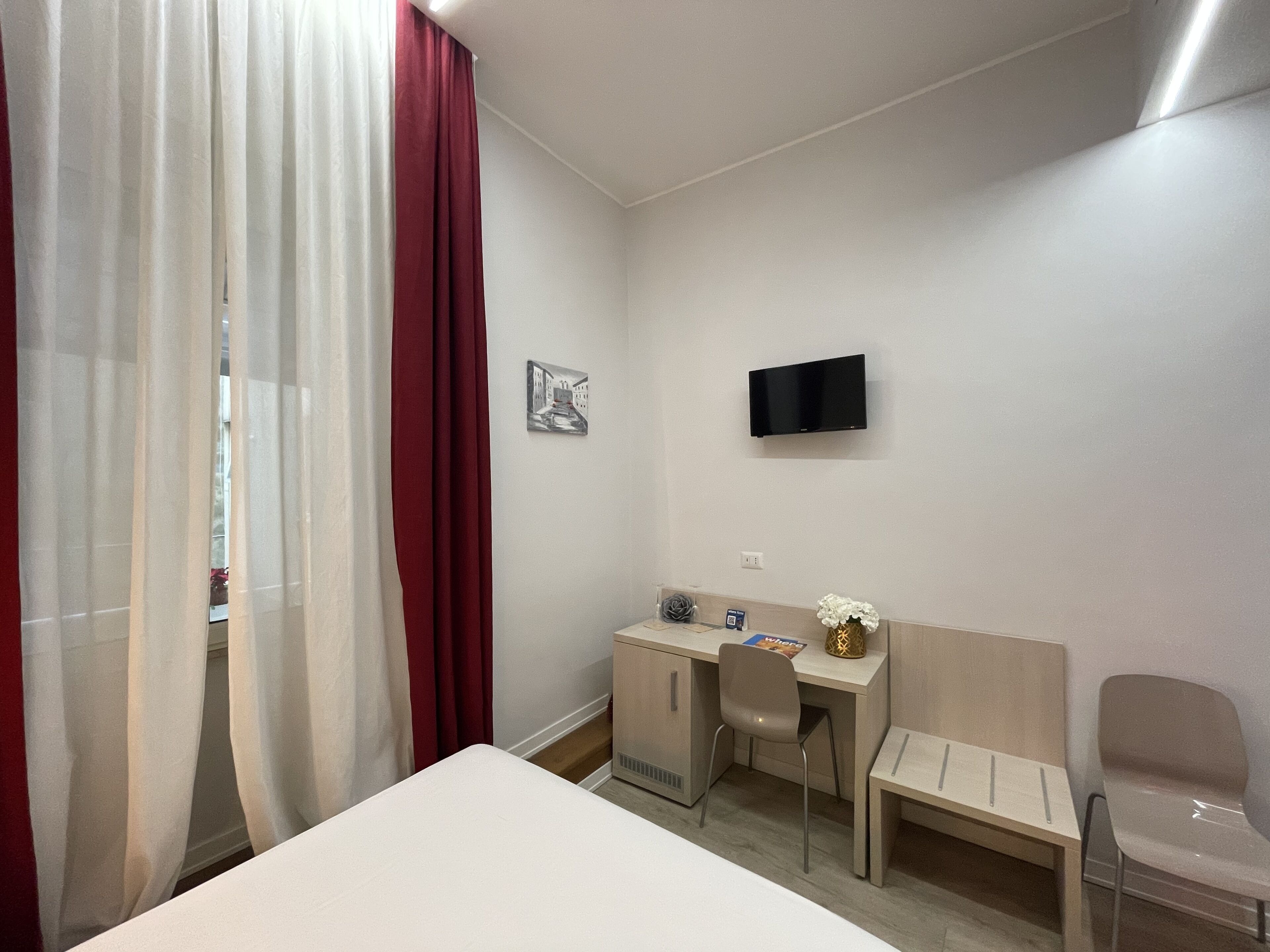 A hotel room with a bed, a desk, a chair, a TV mounted on the wall, and a window with sheer curtains.