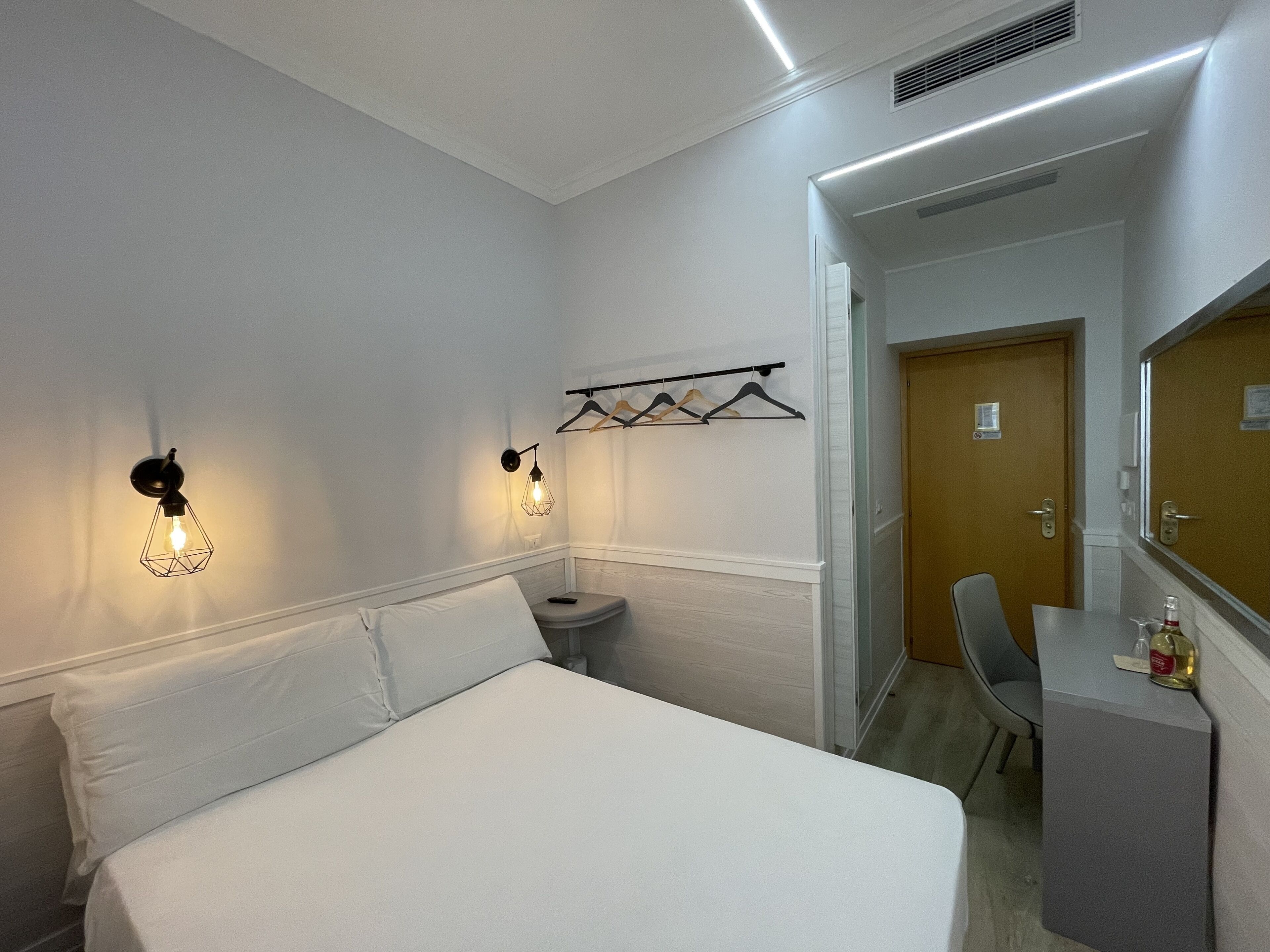 A hotel room with a bed, a desk, a chair, a coat rack, and a door.