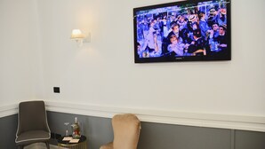 Una habitación con una lámpara de pared, un televisor de pantalla plana montado en la pared, una silla gris, una mesa redonda pequeña y una botella con un vaso.
