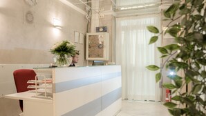 Un mostrador de recepción con una computadora, un reloj y una planta. Se ve una silla roja y un extintor de incendios al fondo.