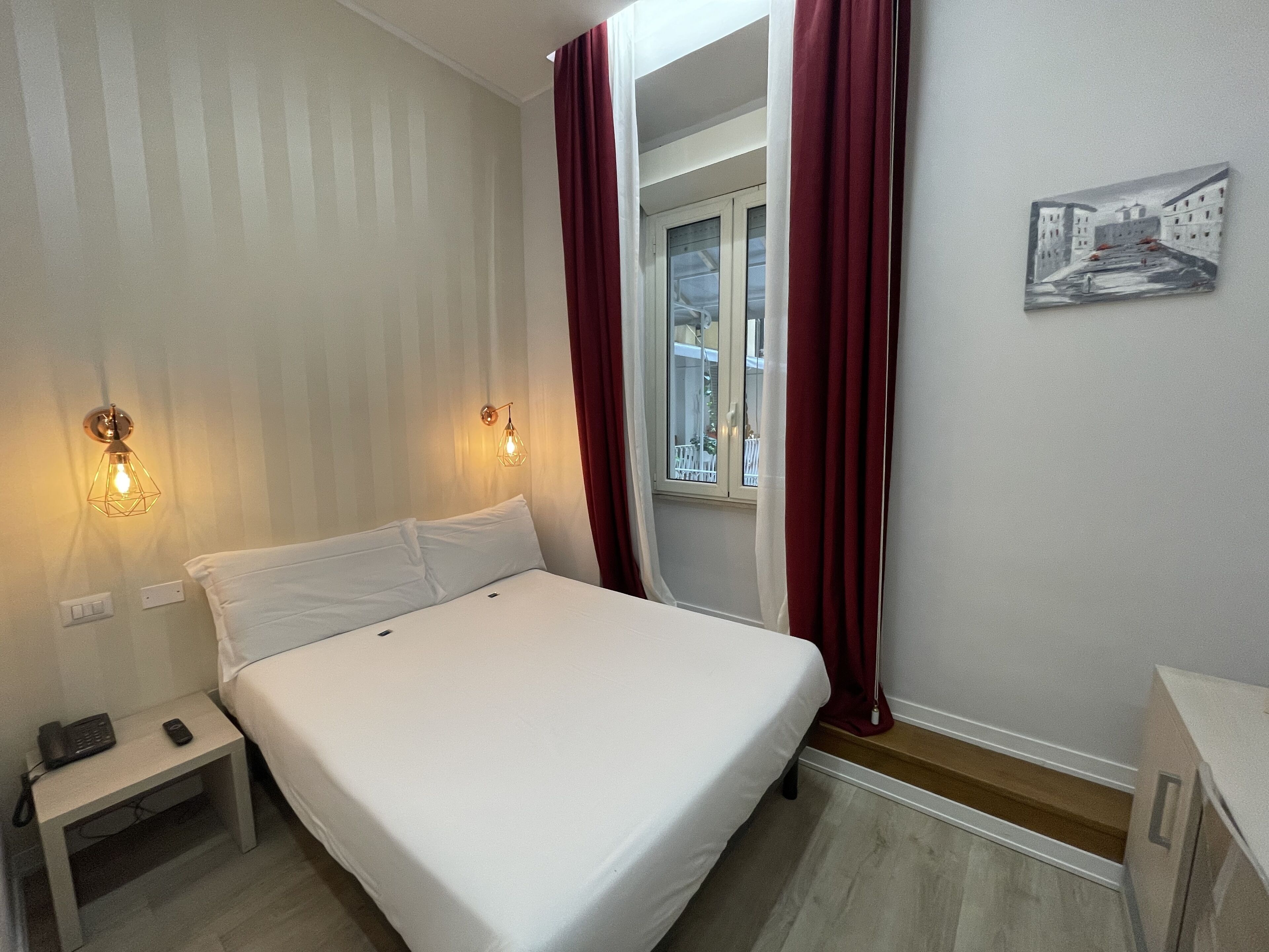 Habitación de hotel con cama, mesita de noche, lámpara de pared, ventana con cortinas y un cuadro en la pared.