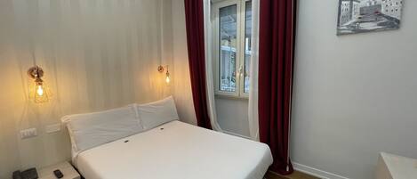 Habitación de hotel con cama, mesita de noche, lámpara de pared, ventana con cortinas y un cuadro en la pared.