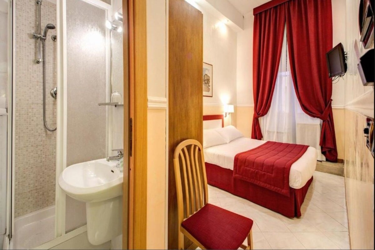 A hotel room with a shower, sink, bed, and a TV mounted on the wall.