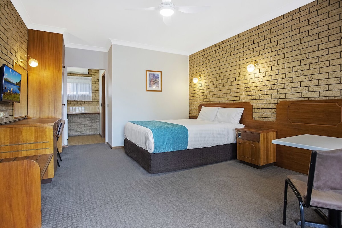 A hotel room with a bed, a desk, a chair, a TV, and a brick wall.