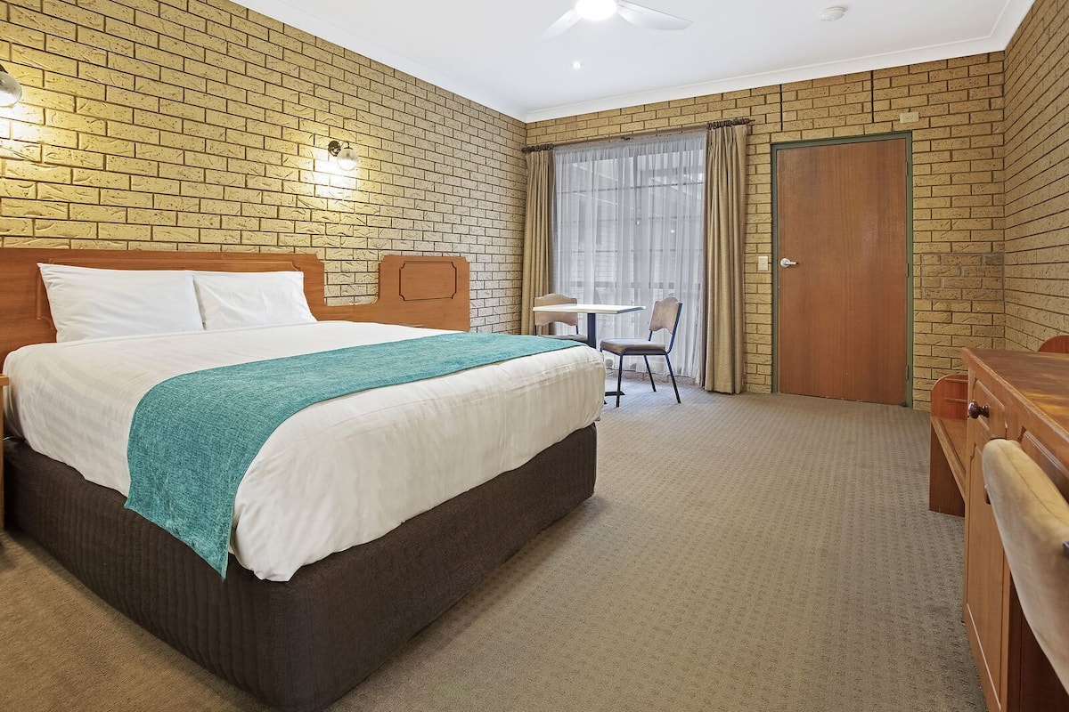 A hotel room with a brick-patterned wall, a bed with white bedding and a teal throw, a wooden desk, a chair, and a window with curtains.