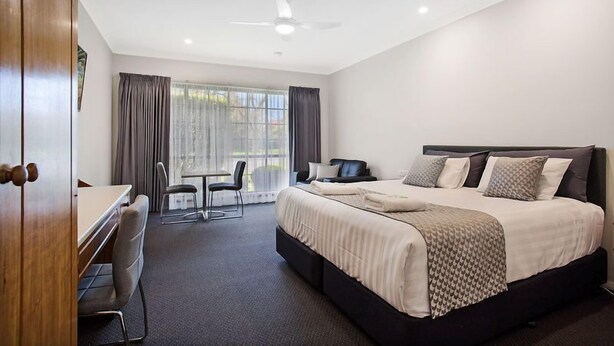 Habitación de hotel con una cama grande, un escritorio y una silla. Hay una ventana con cortinas, un ventilador de techo y vistas al exterior.