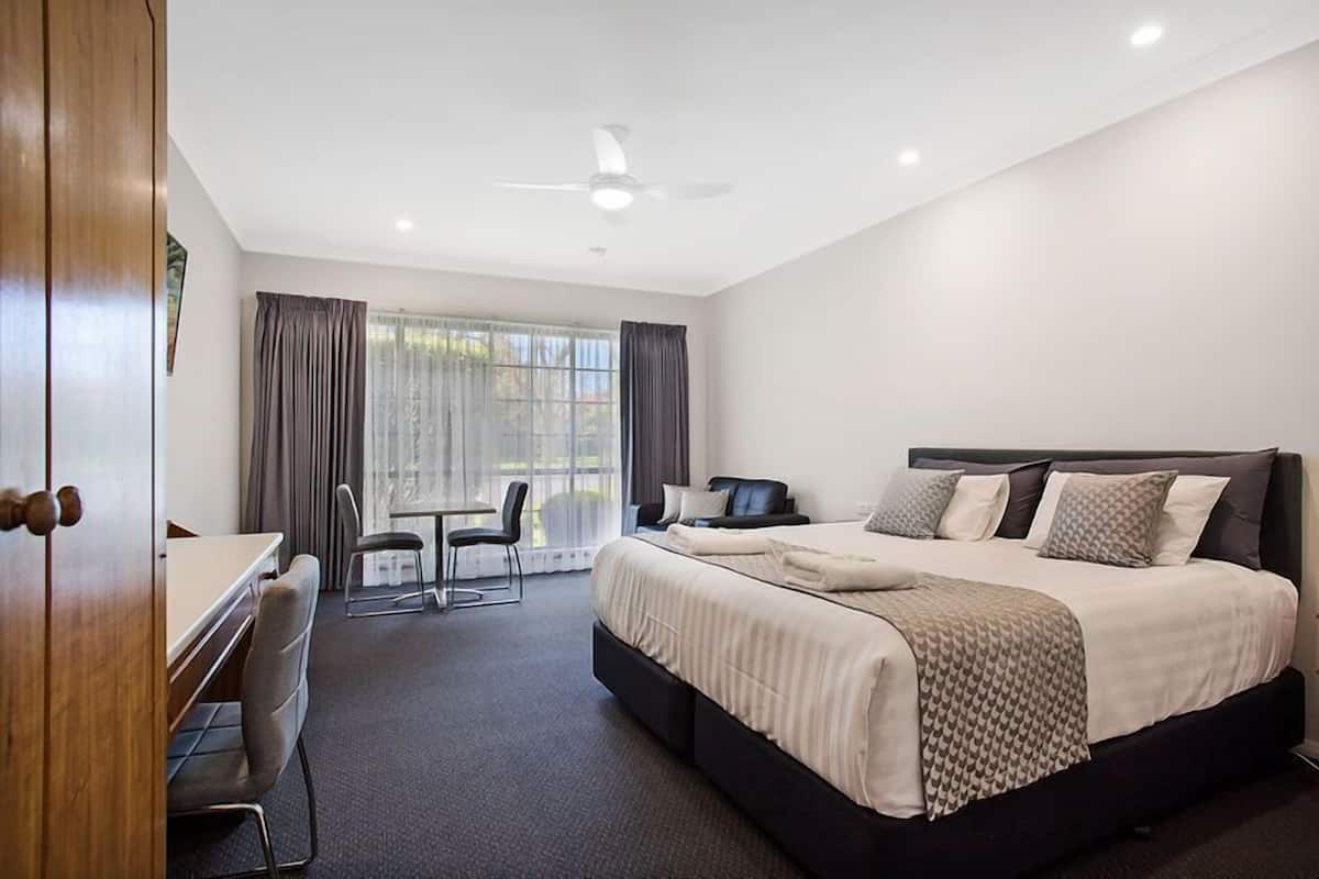 A hotel room with a large bed, a desk, and a chair. There is a window with curtains, a ceiling fan, and a view of the outdoors.