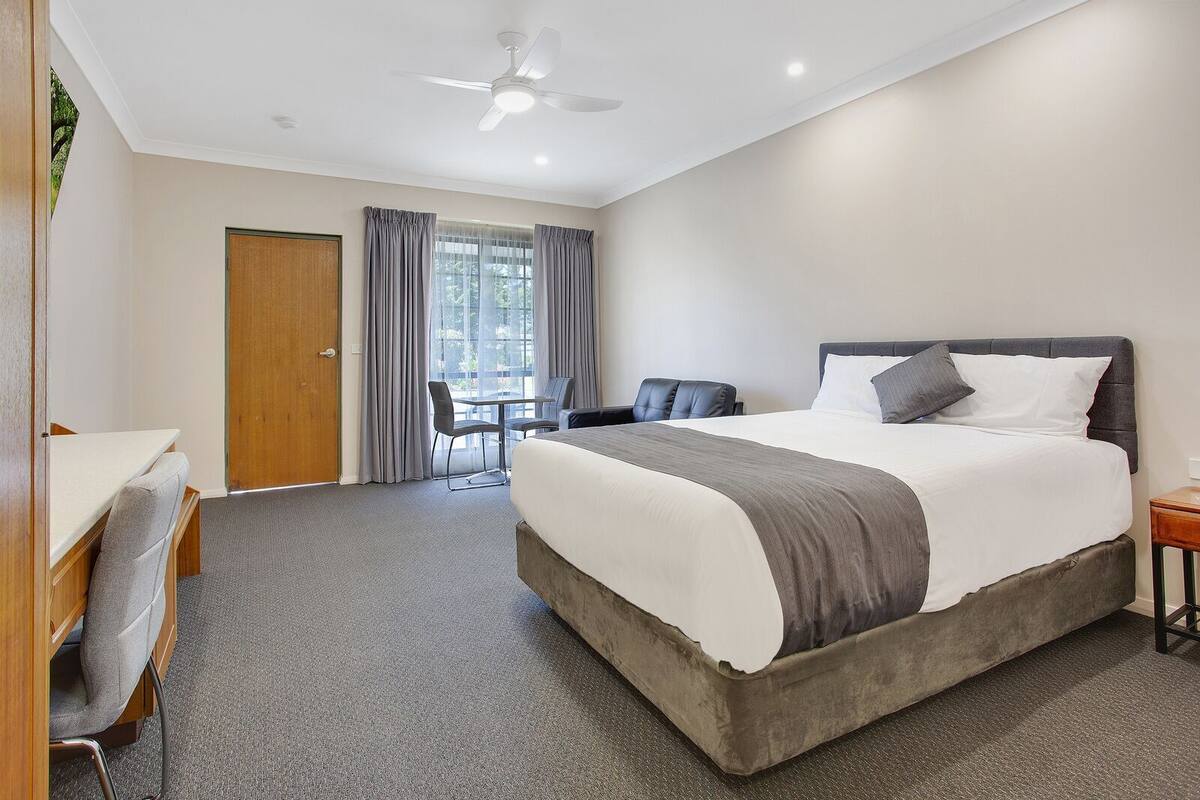A hotel room with a bed, a desk, a chair, a small table, a ceiling fan, and a window with curtains.