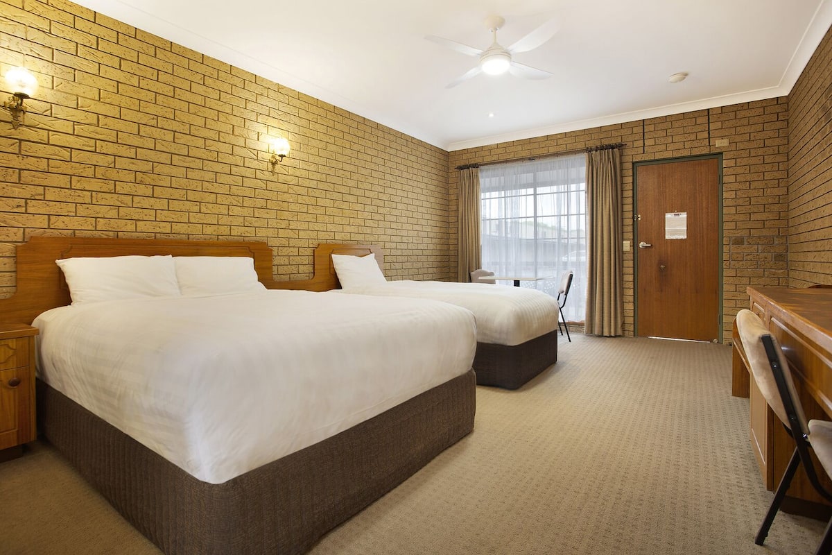 A hotel room with two beds, a desk, a chair, a ceiling fan, and a window with curtains.
