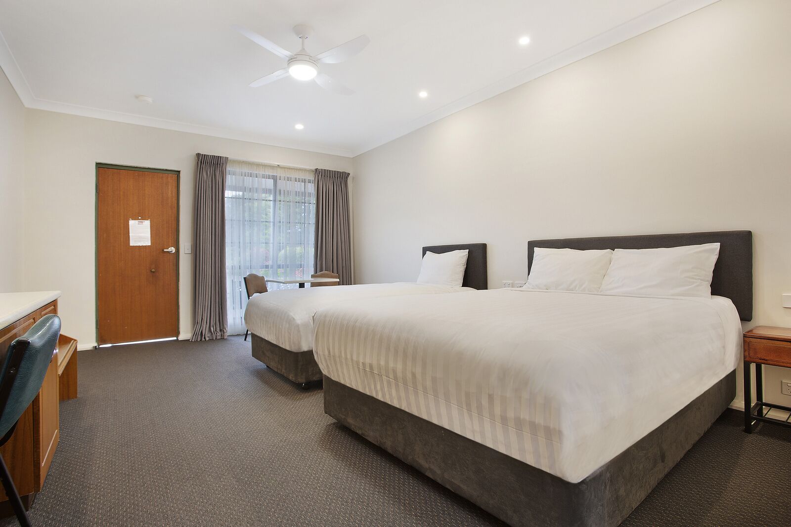 A hotel room with two beds, a desk, a chair, and a ceiling fan.