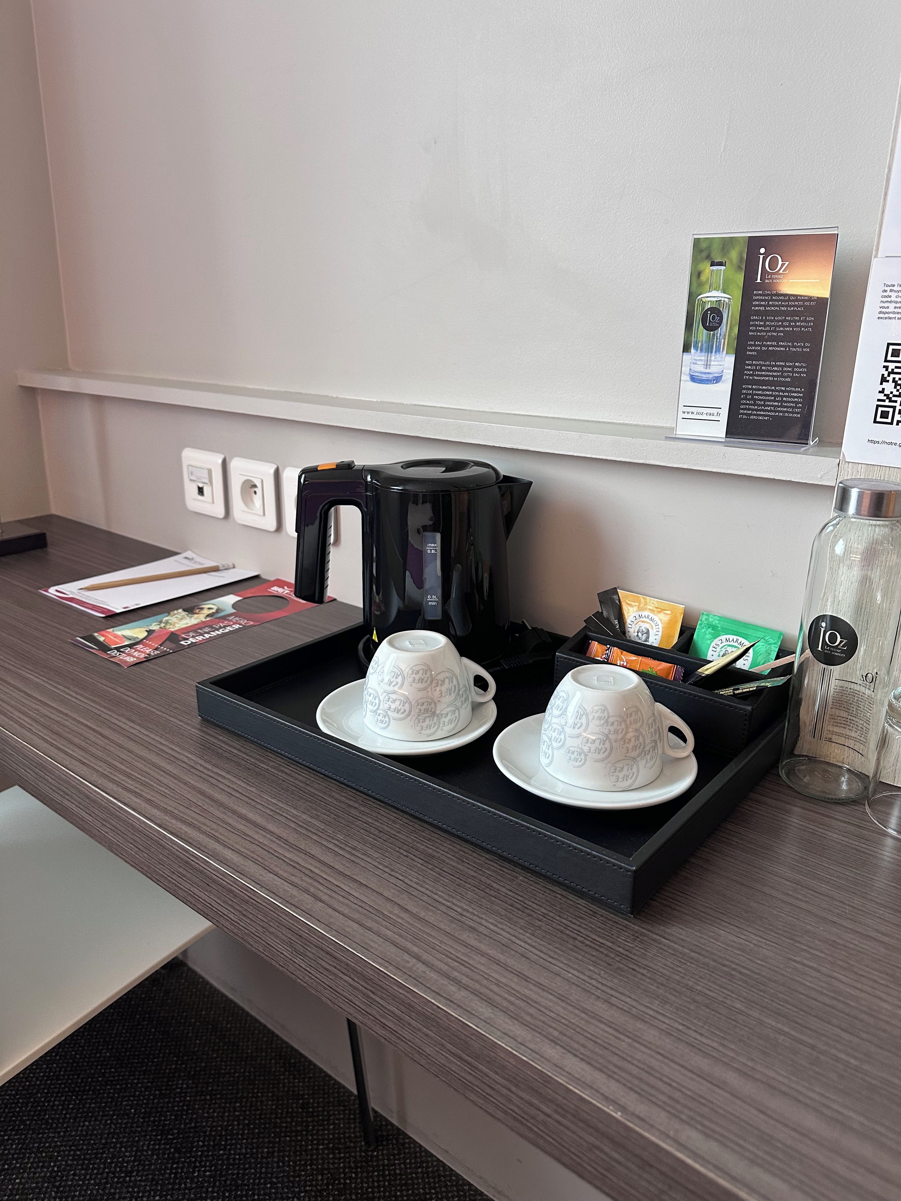 Habitación de hotel con estación de café que incluye una cafetera negra, dos tazas blancas con tapa, una bandeja y un tarro de cristal.