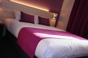 Een net opgemaakt bed met witte en paarse beddengoed, een houten hoofdbord en een nachtkastje met een telefoon.