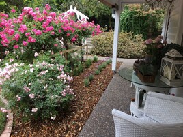 Un jardín con rosas rosadas y blancas, un pérgola blanca y una mesa de vidrio con sillas de mimbre.
