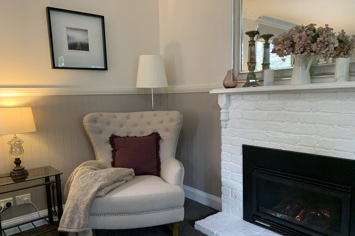 A cozy living room with a white armchair, a fireplace, a framed picture on the wall, and a lamp on a side table.