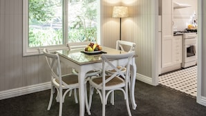 A dining area with a glass table, white chairs, a fruit bowl, and a view of greenery through large windows. - Clarendon Cottages (Sassafras)