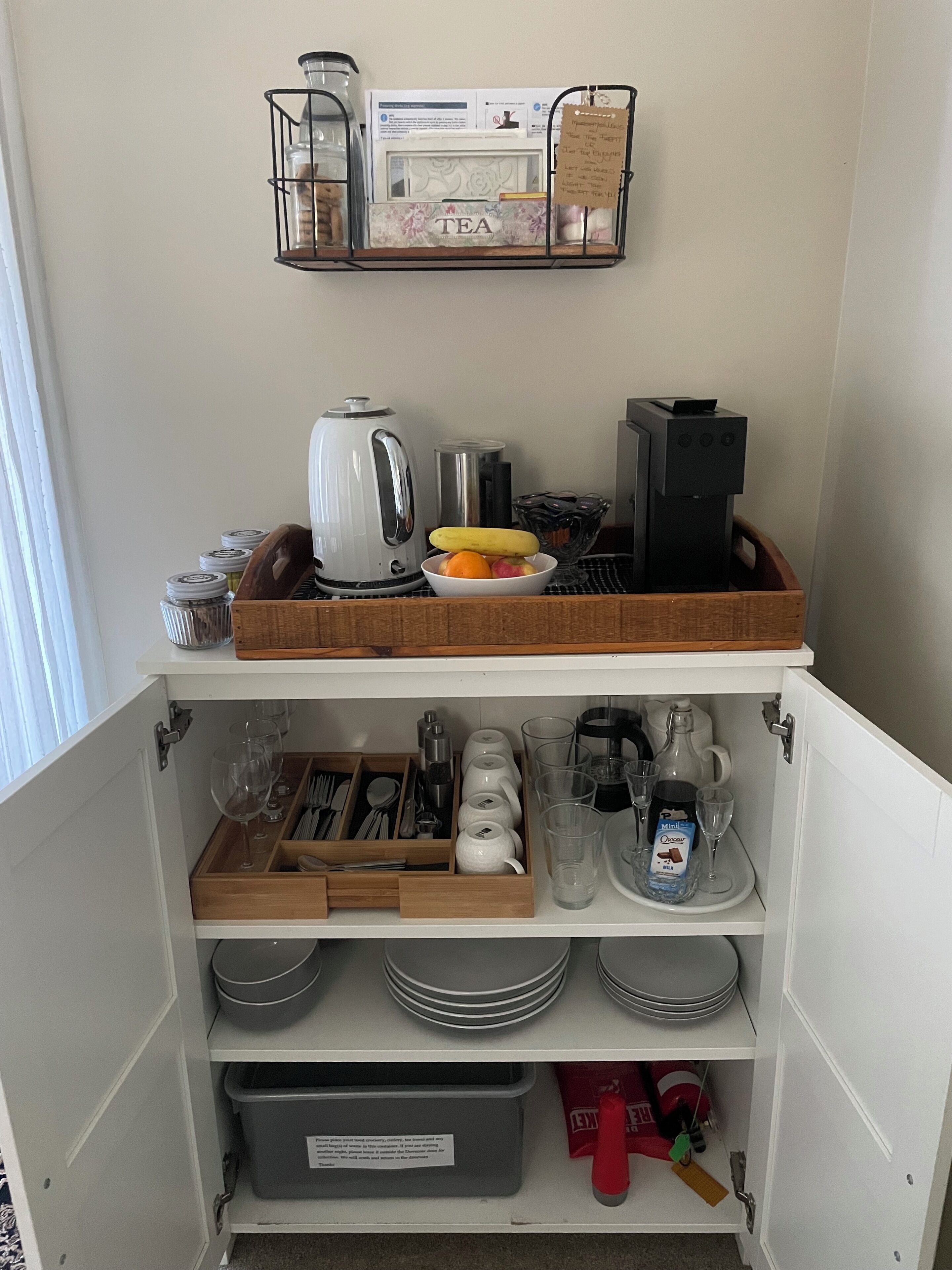 Una cocina con cafetera, hervidor y juego de té sobre la encimera. También hay diversos utensilios de cocina y vajilla guardados en los armarios inferiores.