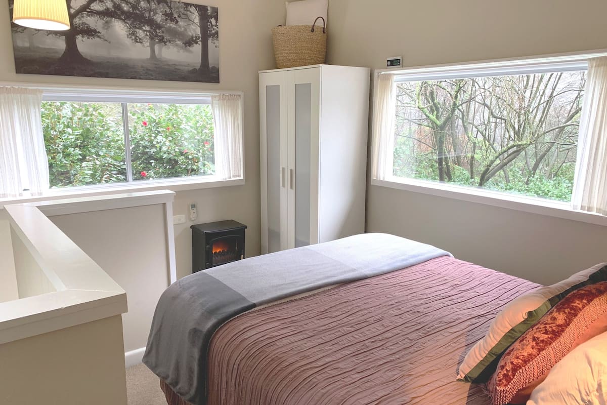 A bedroom with a bed, a heater, a wardrobe, and two windows with views of greenery.