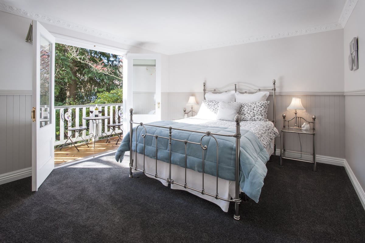 A bedroom with a bed, two bedside tables, a balcony with a railing, and a view of greenery.