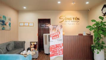 A reception area with a wooden reception desk, a blue sofa, a grey sofa, a wall with a massage service sign, and a 'Super Sale' poster.