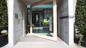 A modern hotel entrance with a glass door, a sign indicating an allergy-friendly environment, and a visible interior with a seating area and a ceiling fan. - NU Hotel (Milan)