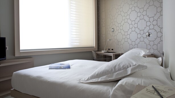 A modern bedroom with a large bed, a desk, and a window with blinds. - NU Hotel (Milan)