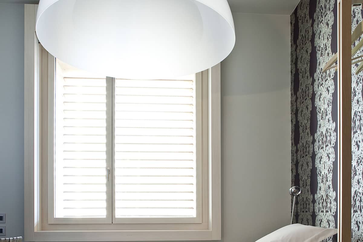 A minimalist bedroom with a bed, a large window with blinds, and a hanging light fixture.