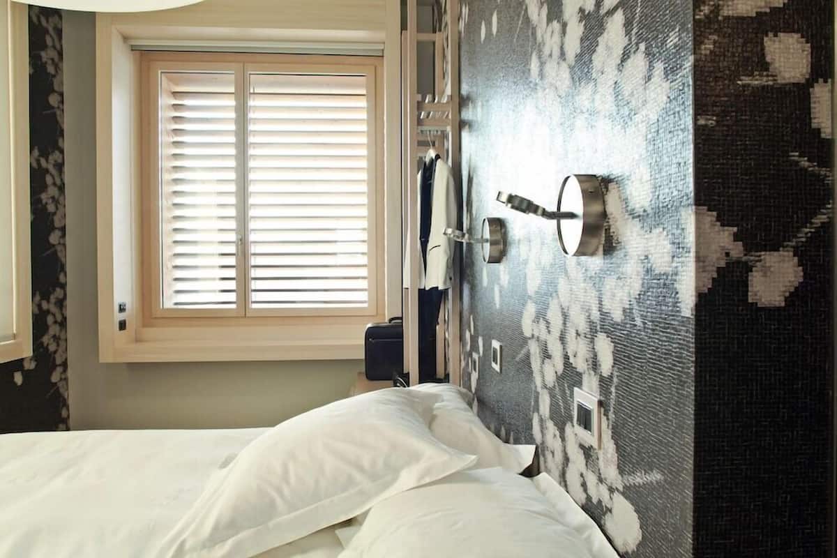 A bedroom with a bed, a window with blinds, a wall-mounted light fixture, and a patterned wall.