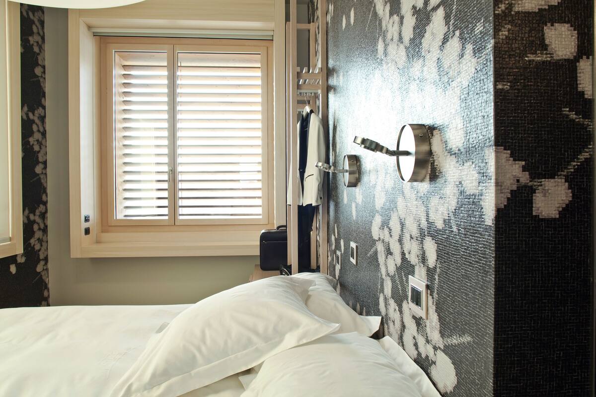 A bedroom with a bed, a window with blinds, a wall-mounted towel rack, and a suitcase on the floor.
