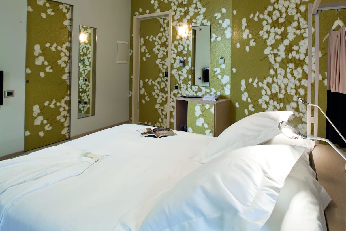 A hotel room with a bed, a bedside table, a mirror, and a hanging garment.