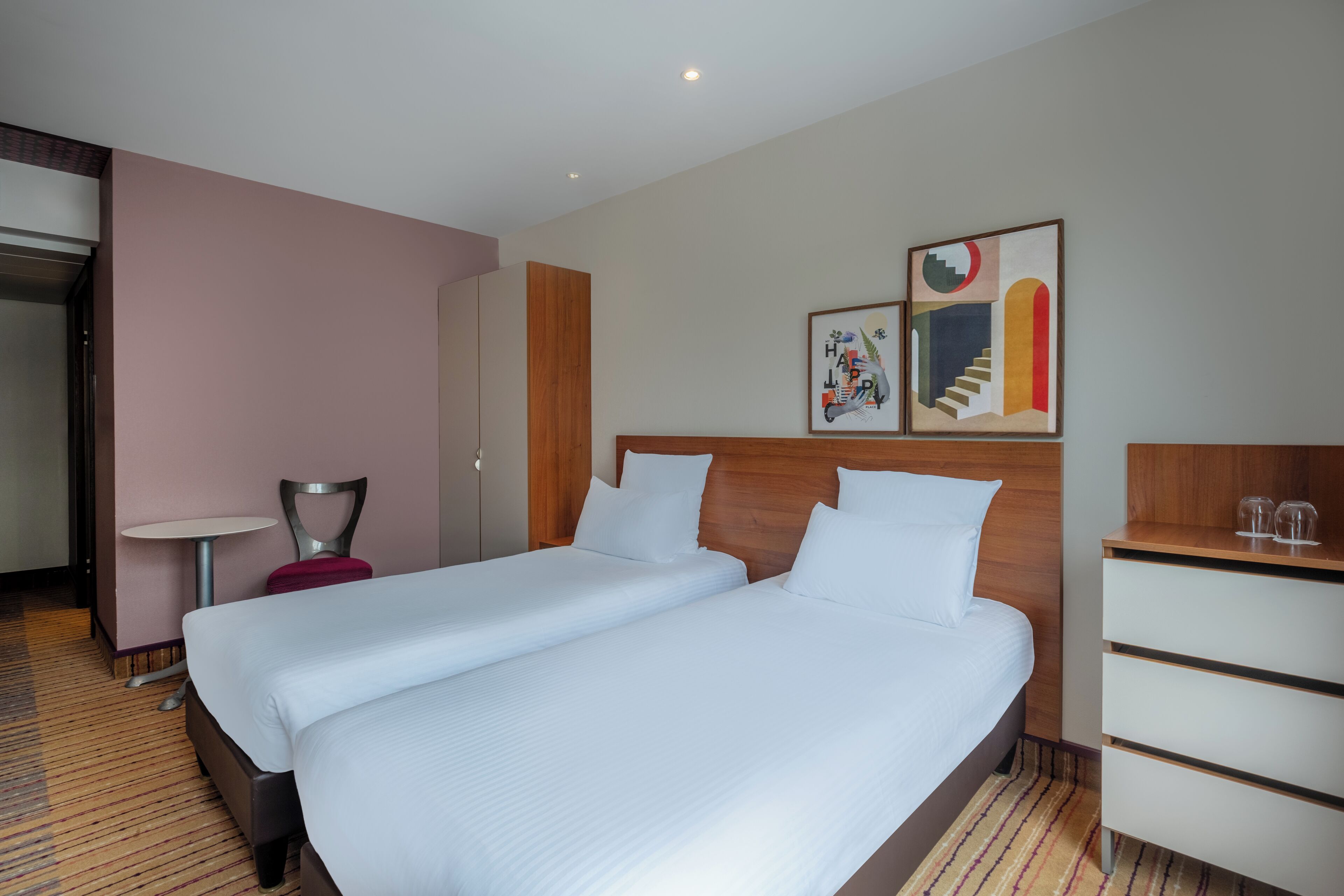 Une chambre d’hôtel avec deux lits, une petite table, une chaise et des œuvres d’art accrochées au mur.