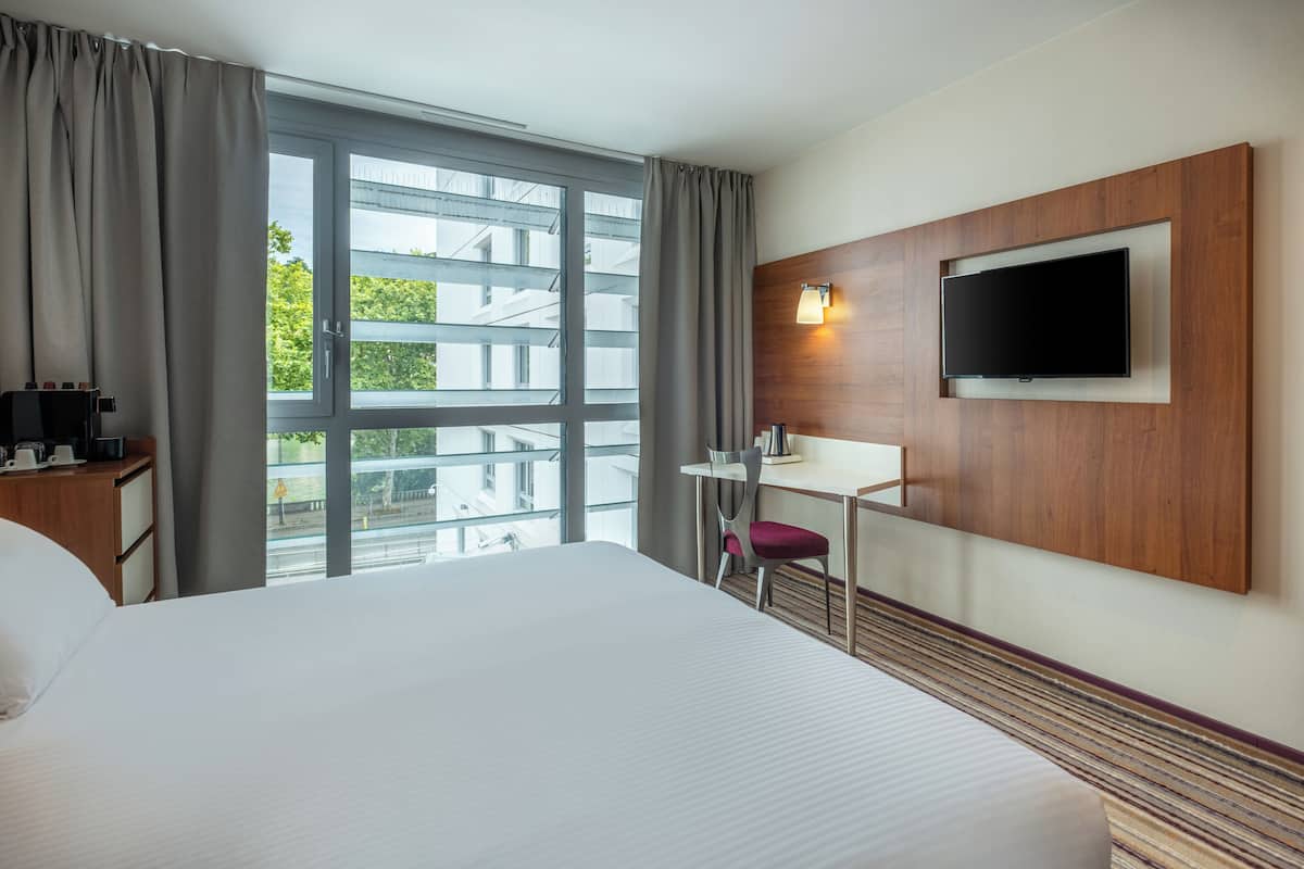 Ein Hotelzimmer mit einem großen Bett, einem Schreibtisch, einem Sessel, einem an der Wand montierten Fernseher und einem Fenster mit Vorhängen.