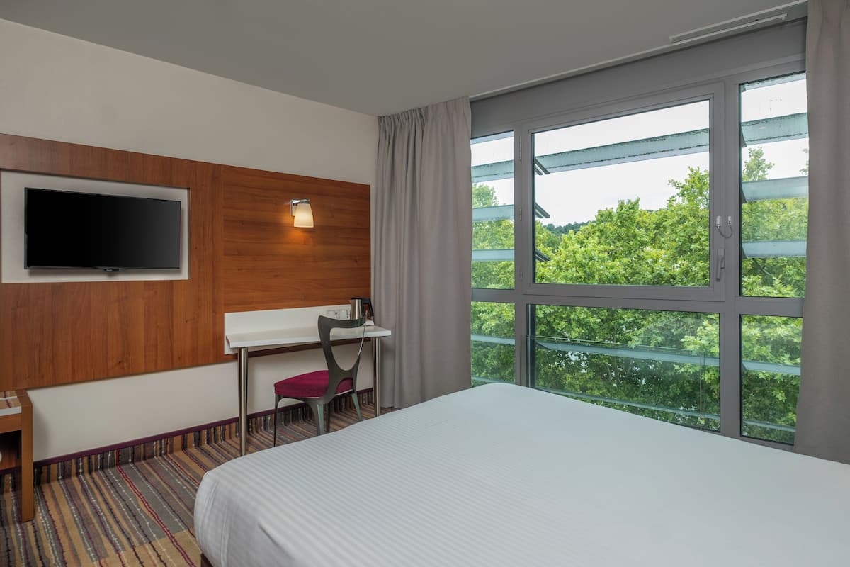 Ein Hotelzimmer mit einem Bett, einem Schreibtisch, einem Sessel, einem Fernseher und einem großen Fenster mit Blick auf Bäume.