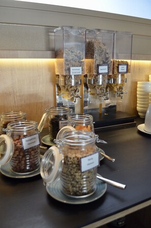 A breakfast bar with various food dispensers and labeled containers of granola and other cereals. - Spark by Hilton Lyon Ouest (Lyon)