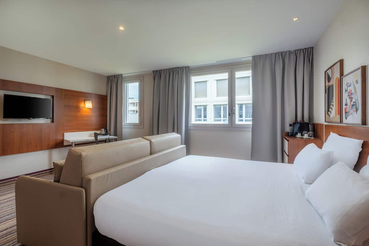 Ein Hotelzimmer mit einem großen Bett, einem an der Wand befestigten Flachbildfernseher, einem Schreibtisch mit Stuhl und einem Fenster mit Vorhängen.