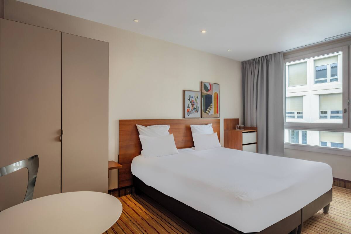 Ein Hotelzimmer mit einem großen Bett, einem hölzernen Kopfteil, einem Nachttisch mit Lampe, einem Sessel und einem Fenster mit Vorhängen.