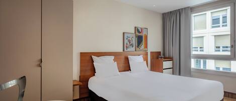 Habitación de hotel con una cama grande, cabecero de madera, mesita de noche con lámpara, una silla y ventana con cortinas.