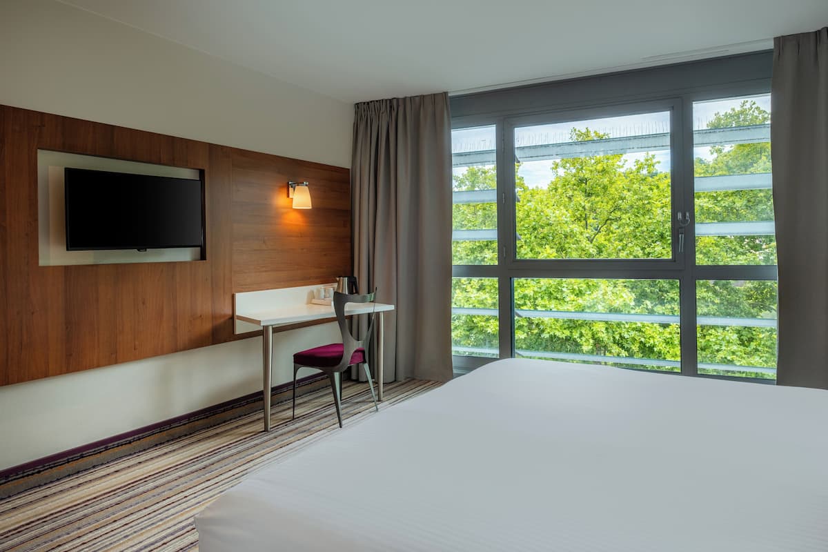 Ein Hotelzimmer mit Bett, Schreibtisch, Stuhl, Wandfernseher und einem großen Fenster mit Blick auf Grünflächen.