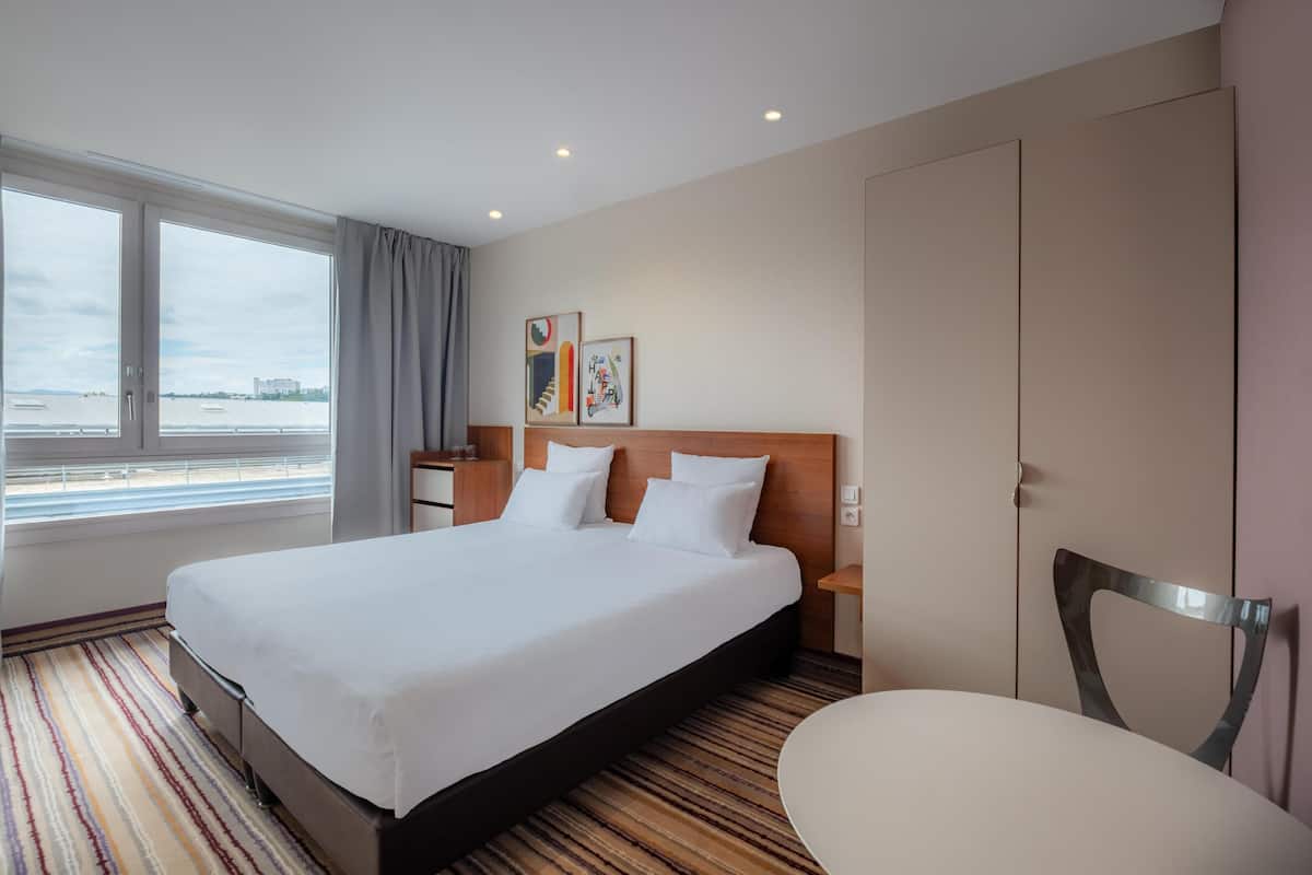 Ein Hotelzimmer mit einem großen Bett, einem Sessel, einem Schreibtisch und einem Fenster mit Blick aufs Wasser.